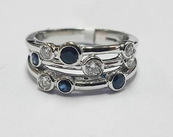 May include: A silver ring with three bands, each adorned with alternating round, bezel-set gemstones. The gemstones include clear diamonds and dark blue sapphires, creating a sparkling and elegant design. The ring is set against a white background.