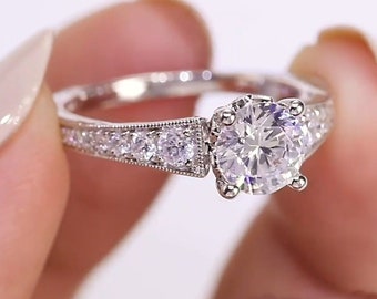 May include: A sparkling diamond engagement ring with a large, round-cut center stone. The ring features a detailed band with smaller accent diamonds. The ring is held up by a person's fingers.