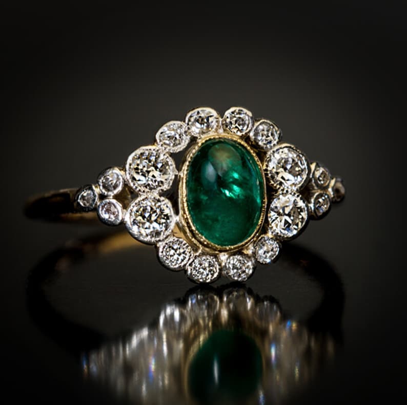 May include: A gold ring featuring an oval, emerald-green gemstone surrounded by a halo of round, sparkling diamonds. The ring's band is thin and delicate, with a classic, elegant design. The diamonds are set in a cluster around the central stone.