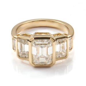 May include: A gold ring with five emerald-cut gemstones. The central stone is the largest, with two smaller stones on either side. The ring has a polished gold band and a classic, elegant design.