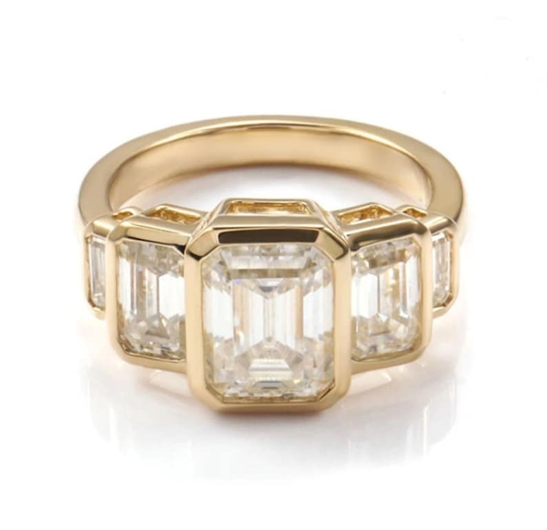 May include: A gold ring with five emerald-cut gemstones. The central stone is the largest, with two smaller stones on either side. The ring has a polished gold band and a classic, elegant design.