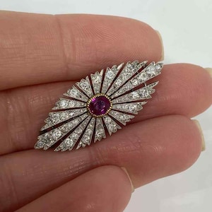 May include: An oval-shaped brooch with a central ruby surrounded by a gold setting. The brooch is further embellished with numerous small diamonds arranged in a sunburst pattern, set in a silver-colored metal.