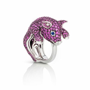 Puede incluir: Anillo de plata con diseño de cerdo. El cerdo está cubierto de piedras preciosas rosas y un ojo de piedra preciosa azul. El anillo tiene un acabado pulido y está sobre un fondo blanco.