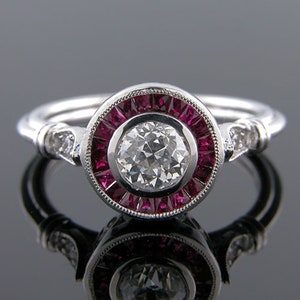 May include: A silver ring with a central, round, clear diamond surrounded by a halo of small, rectangular, deep red gemstones. The ring's band has small, clear accent stones on each side of the central design.