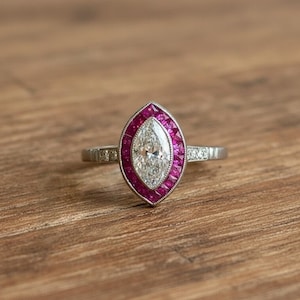 Op de afbeelding: Een zilveren ring met een marquise-vormige middensteen, omringd door een halo van kleine, levendige rode stenen. De ringband is ook versierd met kleinere, heldere stenen. De ring ligt op een warme, houten ondergrond.