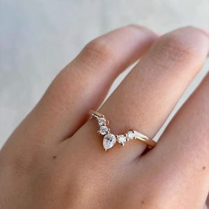 May include: A gold-colored ring with a curved design, set with multiple clear gemstones. The ring features a pear-shaped stone at the center, surrounded by smaller, round stones. The ring is shown on a person's finger.
