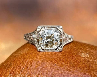 Könnte beinhalten: Ein Verlobungsring im Vintage-Stil mit einem großen, klaren, quadratisch geschliffenen Diamanten, der in einem filigranen Silberband gefasst ist. Der Ring wird auf einer strukturierten, braunen Oberfläche präsentiert, die sein kompliziertes Design und Funkeln hervorhebt.