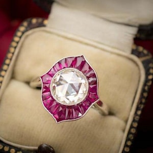 Peut inclure: Gros plan sur une bague de style vintage avec une grosse pierre centrale ronde et transparente. La pierre centrale est entourée d'un halo de petites pierres rouges vives. La bague est sertie dans un anneau argenté et présentée dans un écrin doublé de velours.