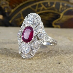 May include: A vintage-style ring featuring a large oval ruby center stone, surrounded by smaller diamonds. The ring has a detailed, ornate design with a silver band. The ruby is a deep red color, and the diamonds sparkle against the silver.