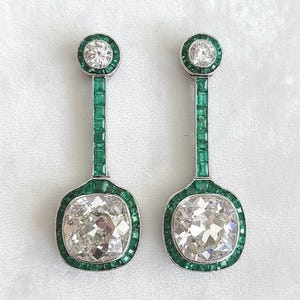 Op de afbeelding: Een paar elegante oorhangers met grote, sprankelende diamanten met kussenslijpsel. De diamanten zijn omlijst door een rand van smaragdgroene edelstenen en geaccentueerd met kleinere diamanten en groene edelstenen. De oorbellen hebben een zilverkleurige metalen zetting.