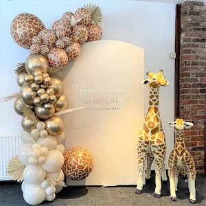 Puede incluir: Un telón de fondo blanco con el texto "Rosie Olivia  CHRISTENING 13th October 2021" está decorado con un arco de globos en oro, blanco y marrón con estampado de jirafa. Dos jirafas de peluche se encuentran frente al telón de fondo.