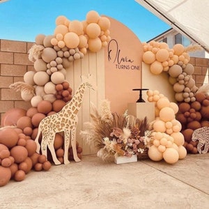 May include: A wooden giraffe cutout stands in front of a backdrop decorated with beige and brown balloons. The backdrop also features a peach-coloured semi-circle with the text "Olivia Turns One" in brown script.