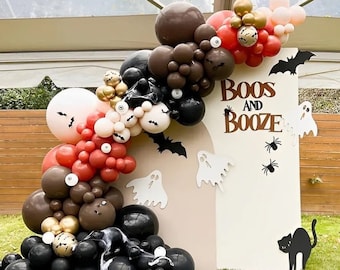 May include: A Halloween party backdrop with a white sign that says "Boos and Booze". The sign is decorated with black bats, spiders, and ghosts. A balloon arch made of black, brown, red, and white balloons is in front of the sign.