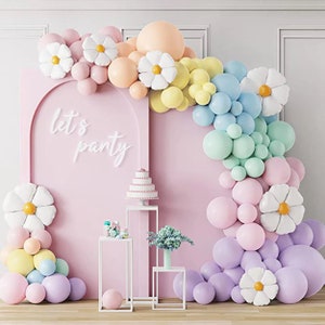 以下が含まれることがあります： パステルピンクの背景に、「let's party」という白い文字が書かれています。パステルカラーの風船と白いデイジー型の装飾でできた風船アーチ。アーチは画像の右側です。ケーキが置かれた一番上のスタンドと、他の装飾が置かれた2つのスタンドを含む、白い金属製のスタンドが3つあります。