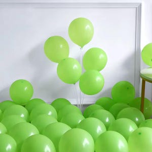May include: A display of green balloons, with some scattered on the floor and others held up by a clear stand. The balloons are a vibrant, solid green, giving a celebratory feel. The backdrop is a plain white wall.