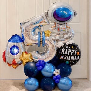 May include: A festive balloon arrangement featuring a silver number 5, an astronaut, and a rocket ship. The arrangement includes blue and silver balloons, gold star accents, and a black balloon with "Happy Birthday" text. Perfect for a space-themed birthday.