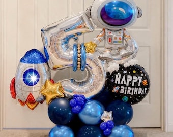 May include: A festive balloon arrangement featuring a silver number 5, an astronaut, and a rocket ship. The arrangement includes blue and silver balloons, gold star accents, and a black balloon with "Happy Birthday" text. Perfect for a space-themed birthday.