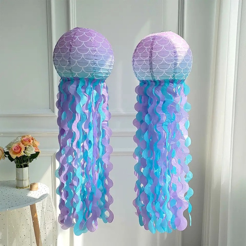 Op de afbeelding: Twee paarse en blauwe ombre-papierlanterns met een zeemeermin-schubbenpatroon. De lantaarns hangen aan een plafond en hebben blauwe en paarse tissue-papier streamers aan de onderkant bevestigd.