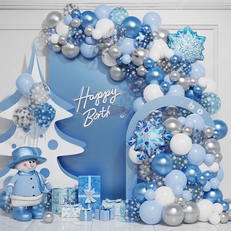 以下が含まれることがあります： 雪だるま、プレゼント、そして「Happy Birth」という銀色のキラキラ文字が書かれた風船アーチのある、青と白の冬のワンダーランドの誕生日パーティーの背景です。