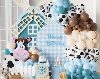 May include: A blue and white checkered backdrop with the words "Happy Birthday" in silver foil letters. A windmill with a cow balloon and a brown and white cow balloon. A balloon arch with blue, brown, white, and black balloons.