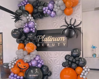 May include: A Halloween balloon arch featuring orange, black, purple, gray, and silver balloons, including a pumpkin-shaped balloon and two spider-shaped balloons, framing a black sign with the white text Platinum BEAUTY.