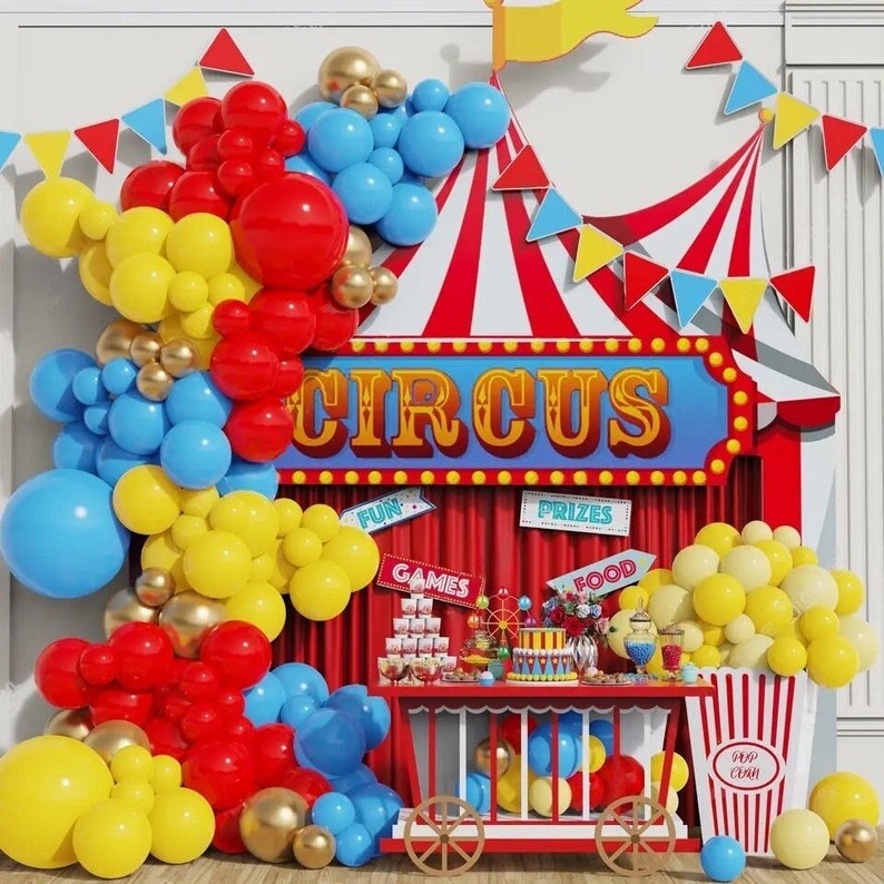 May include: A circus-themed party backdrop with a red and white striped tent, a sign that says "CIRCUS", and a table with various treats and decorations. The backdrop is decorated with red, yellow, and blue balloons.