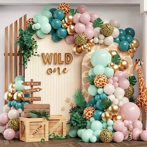 Könnte beinhalten: Eine Dschungel-Motto-Geburtstagsdekoration für einen ersten Geburtstag mit einem Holzzaun, grünen Blättern und einem Ballonbogen in den Farben Türkis, Rosa, Gold und Weiß. Der Bogen ist mit Leopardenmuster-Ballons und Holzschilden verziert, auf denen "Wild One", "Be Brave", "Adventure" und "Wild Animals" steht.