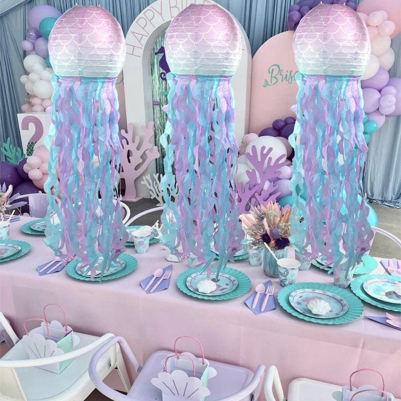 Op de afbeelding: Drie paarse en blauwe decoraties met een kwal-thema met papieren slingers die eronder hangen. De decoraties zijn op een tafel met een roze tafelkleed en witte stoelen geplaatst. De tafel is gedecoreerd met borden, bekers en servetten met een zeemeermin-thema.
