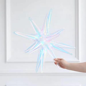 May include: An iridescent star-shaped decoration with multiple points. The star has a holographic, shimmering effect, reflecting various colours. The decoration is held by a person's hand, suggesting it is lightweight and possibly a balloon or ornament.
