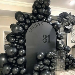 Peut inclure: Une arche de ballons noirs encadre un fond gris foncé avec le nom "LEURYS" et le chiffre "31". La scène comprend un petit gâteau sur un présentoir transparent et des ballons noirs supplémentaires, créant une ambiance festive.