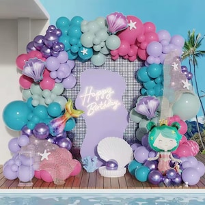 May include: A mermaid-themed birthday party decoration set. The backdrop features a large arrangement of balloons in shades of teal, purple, pink, and white. A sign with "Happy Birthday" is in the center. Mermaid tail and seashell accents complete the festive scene.
