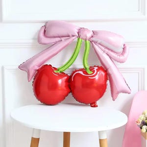 Puede incluir: Globo rojo con forma de cereza con tallos verdes y un lazo rosa. El globo está sobre una mesa blanca. Decoración festiva.