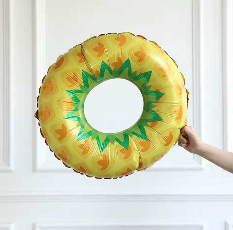 May include: A yellow and green pineapple-shaped inflatable ring. The ring has a large circular opening in the centre and is decorated with a pineapple pattern. The ring is held by a person's hand.