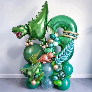 May include: A green dinosaur balloon with a number 8 balloon, surrounded by smaller green, blue, and gold balloons. The balloons are decorated with green leaves and a dinosaur print.