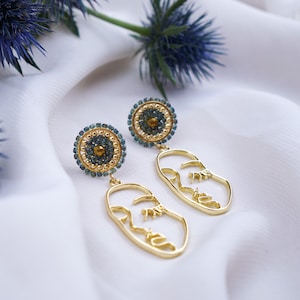 May include: Gold earrings with a blue and gold beaded circle and a gold face outline.