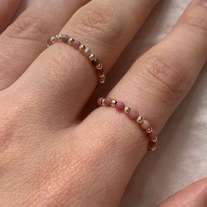 Puede incluir: Dos delicados anillos de cuentas con cuentas rosadas y doradas. Los anillos se llevan en el índice y el dedo medio de una mano.