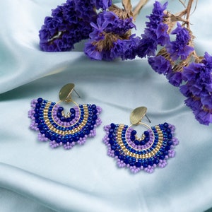 May include: A pair of beaded earrings with a gold hoop and a fan-shaped design. The earrings are made with purple and blue beads and have a gold accent.