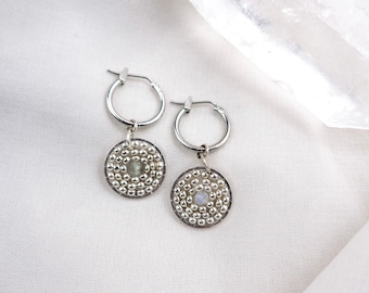 May include: A pair of silver hoop earrings with a small round charm hanging from each hoop. The charms are made of silver beads and feature a small gemstone in the centre. The earrings are displayed on a white fabric background.