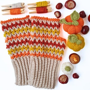 May include: A pair of crocheted fingerless gloves with a chevron pattern in orange, yellow and red. The gloves have a beige cuff and are surrounded by autumn-themed decorations.