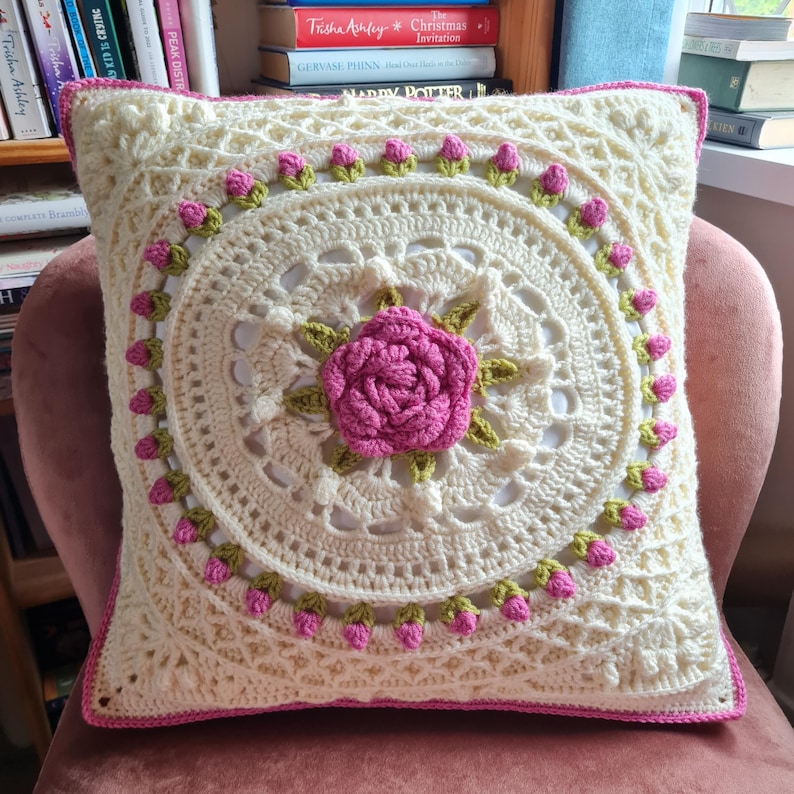 Op de afbeelding: Een gehaakt kussen met een witte achtergrond en een roze roos in het midden. Het kussen is omgeven door een rand van groene en roze gehaakte bloemen.