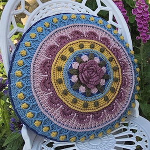 Puede incluir: Un cojín redondo de ganchillo con un diseño floral. El cojín presenta círculos concéntricos en tonos azules, amarillos, rosas y morados. Una gran rosa en 3D está en el centro. El cojín está sobre una silla blanca ornamentada.