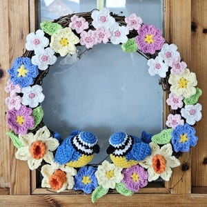 Op de afbeelding: Een gehaakte krans versierd met kleurrijke bloemen en twee blauwe vogels. De krans is gemaakt met verschillende garenkleuren, waaronder roze, geel, wit en blauw. De vogels zitten op de krans en kijken elkaar aan.