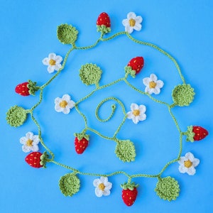 May include: A crocheted garland featuring alternating red strawberries and white flowers. The garland is crafted from yarn and strung together with white thread. It's ideal for adding a touch of charm to a home or party.