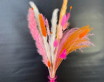 May include: A colourful bouquet of dried flowers in a pink glass vase. The arrangement includes pink, orange, white, and peach-coloured plumes and stems. The vase is a bright pink, and the background is a dark grey.