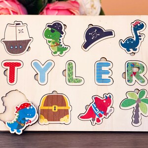 May include: Wooden puzzle with a pirate theme. The puzzle features a ship, a dinosaur, a treasure chest, a palm tree, and the name "TYLER".