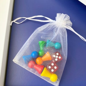 Op de afbeelding: Een witte mesh tas gevuld met kleurrijke houten spelstukken en dobbelstenen. De stukken zijn rood, oranje, geel, groen, blauw en paars. De dobbelstenen zijn rood met witte stippen.