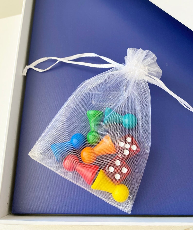 Op de afbeelding: Een witte mesh tas gevuld met kleurrijke houten spelstukken en dobbelstenen. De stukken zijn rood, oranje, geel, groen, blauw en paars. De dobbelstenen zijn rood met witte stippen.