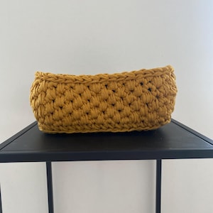 Op de afbeelding: Mosterdgele gehaakte mand. De rechthoekige mand is gemaakt van dik, getextureerd garen. De mand staat op een zwart oppervlak. De mand is een decoratief opbergitem.