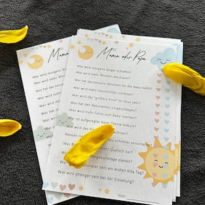 May include: Stack of baby shower game cards with a sun, moon, and cloud design. The cards have questions in German about the parents' roles. Yellow flower petals are scattered around the cards on a dark surface.