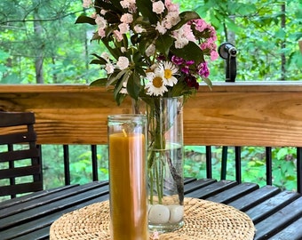 May include: A brown pillar candle in a clear glass holder sits on a woven placemat. A bouquet of pink and white flowers in a clear glass vase is behind the candle.